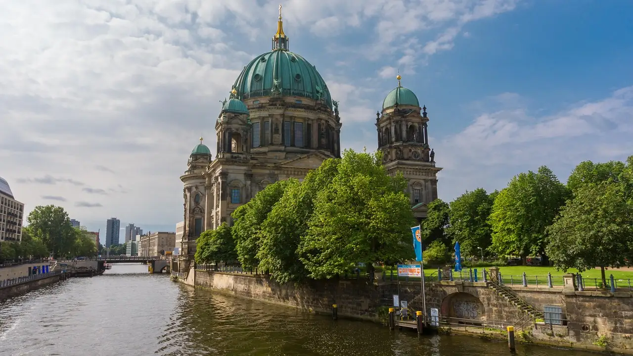 Der Berliner Dom mit seiner grünen Kuppel, gesehen über die Spree hinter belaubten Bäumen an einem bewölkten Tag