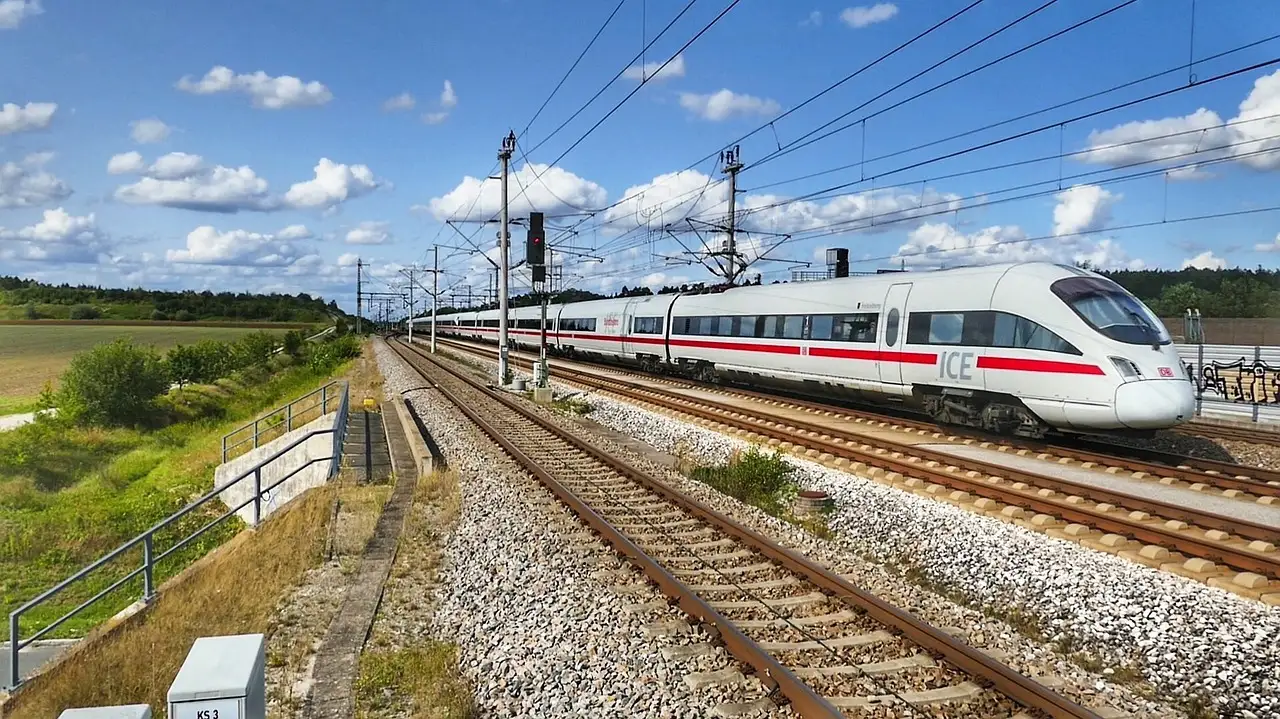 Ein weißer ICE-Hochgeschwindigkeitszug fährt auf einer elektrifizierten, zweigleisigen Strecke unter blauem Himmel