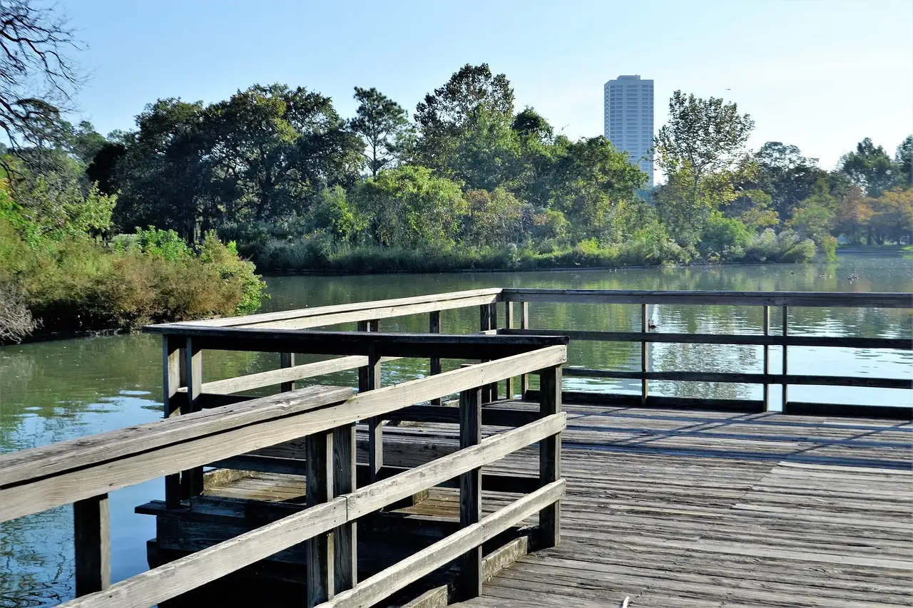 Ein Holzsteg am See im Hermann Park, dahinter Bäume und ein hoher Turm