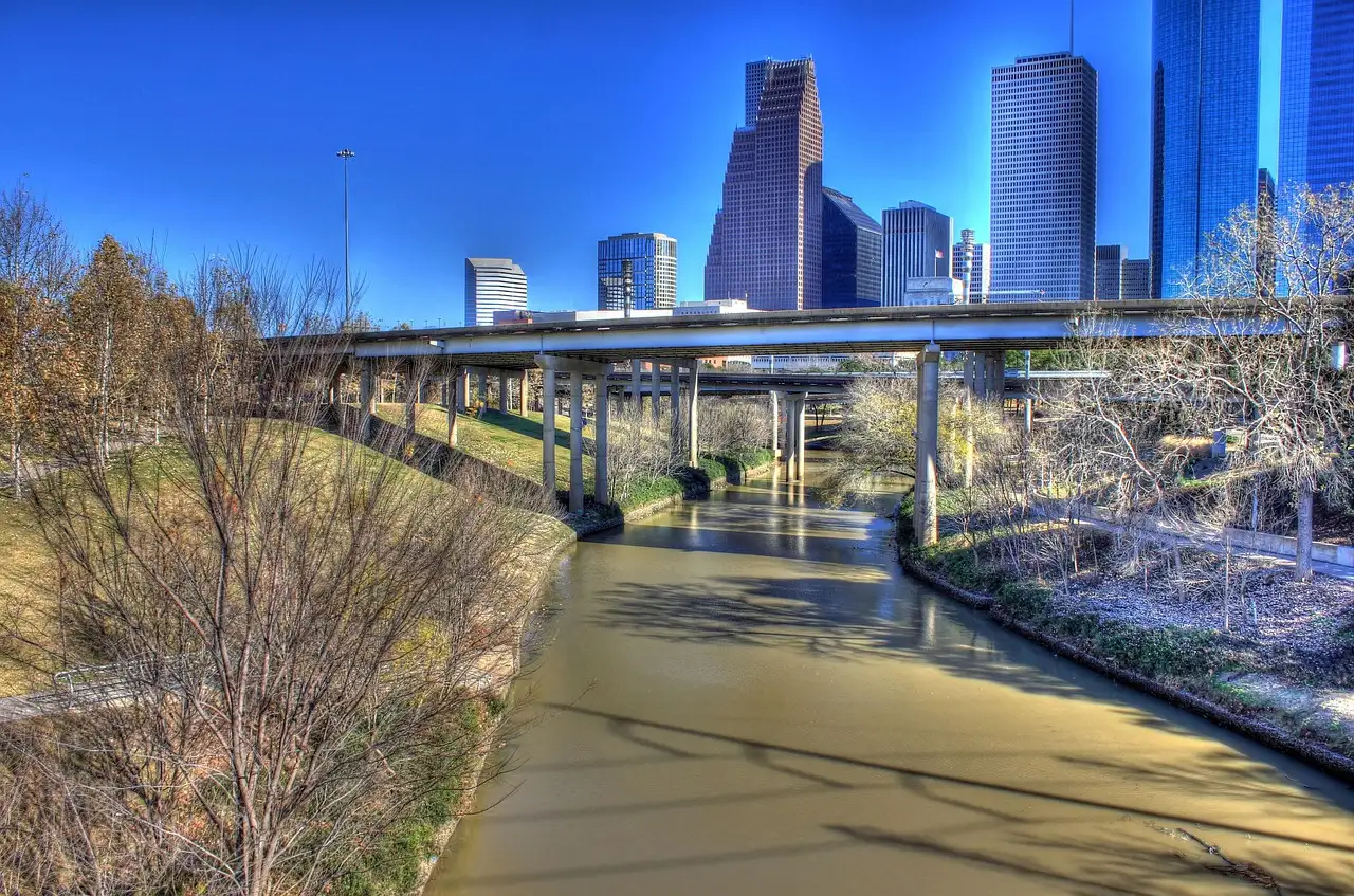 Ein asphaltierter Weg am Bayou mit kahlen Bäumen unter einer Autobahnbrücke, dahinter die Hochhäuser von Houston