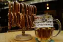 Ein Glaskrug mit bernsteinfarbenem Bier neben einem Holzständer mit Brezen in einer schummrigen Bierhalle