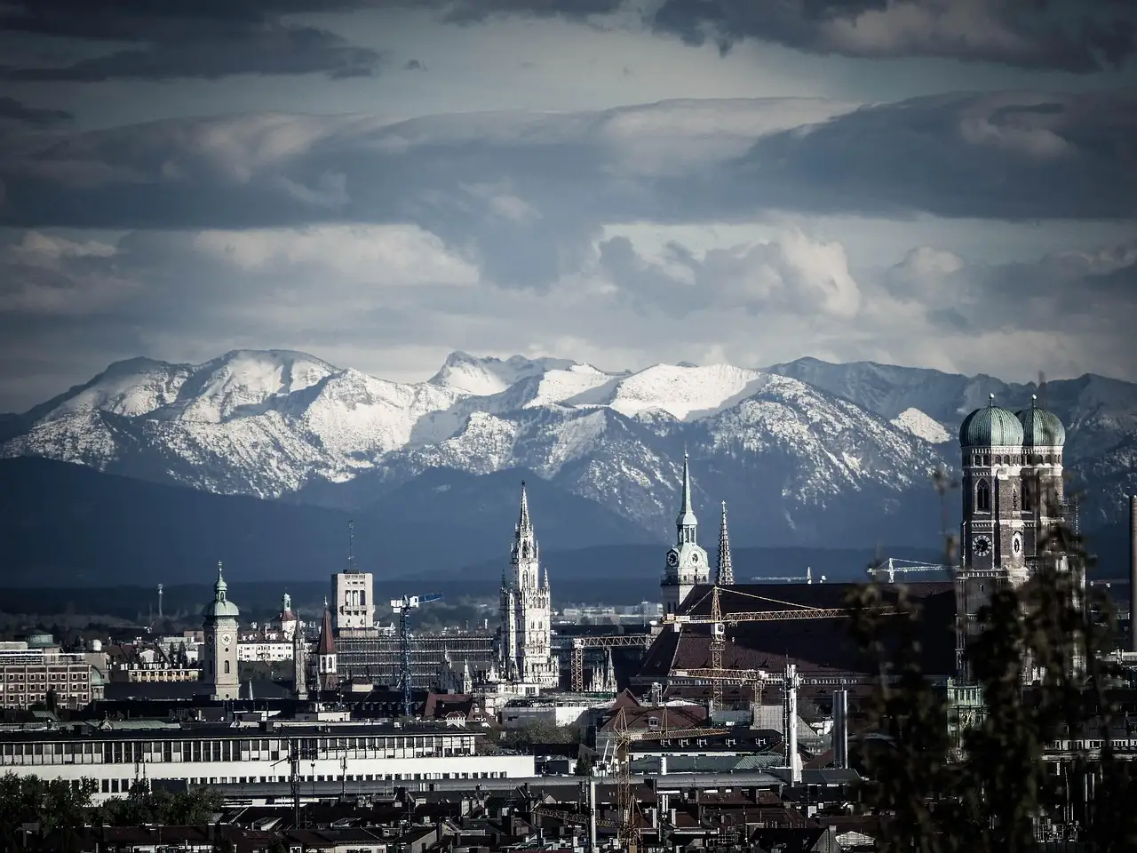 Die Türme der Frauenkirche über der Münchner Altstadt, dahinter die verschneiten Alpen unter dunklen Wolken