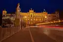 Das Maximilianeum nachts beleuchtet am Ende einer Brücke, davor eine Statue und Lichtspuren von Autos