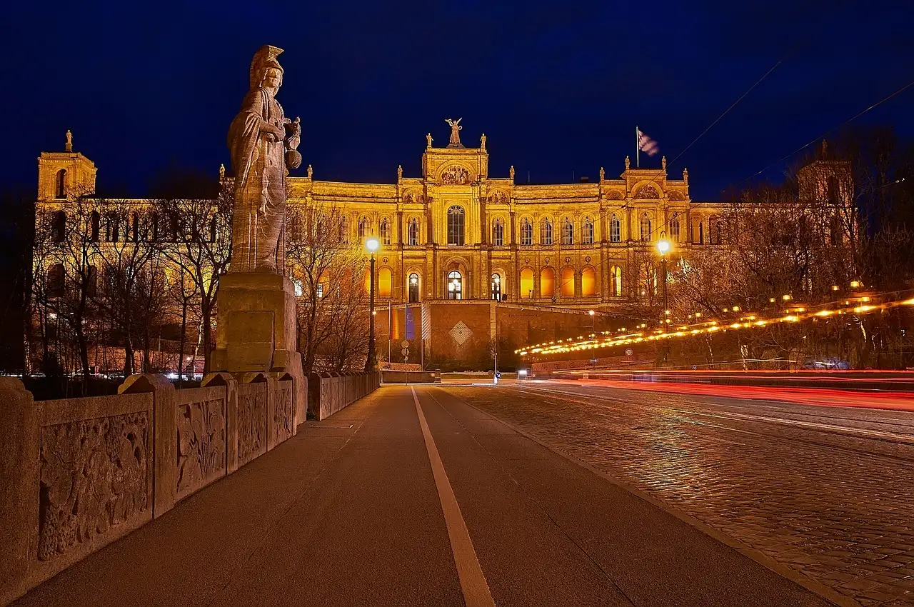 Das Maximilianeum nachts beleuchtet am Ende einer Brücke, davor eine Statue und Lichtspuren von Autos