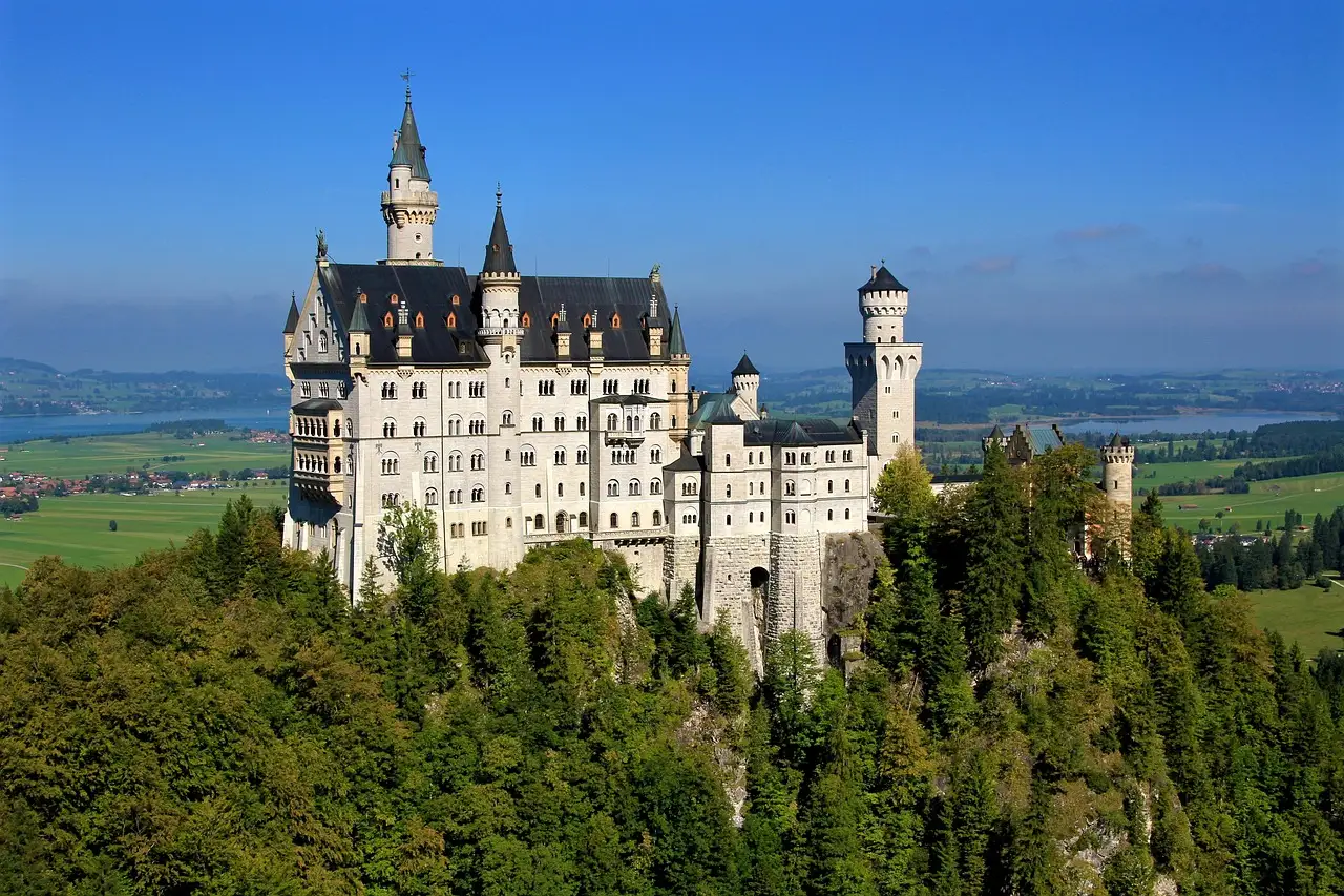 Schloss Neuschwanstein auf seinem Felsen über grünem Wald, dahinter die Ebene und ein See