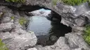 Ein Höhlenbogen aus Kalkstein über klarem Wasser, umgeben von grauem Fels und Farnen