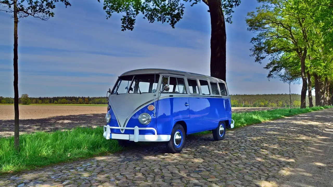 Una furgoneta clásica Volkswagen T1 azul y blanca aparcada en un camino empedrado bajo los árboles