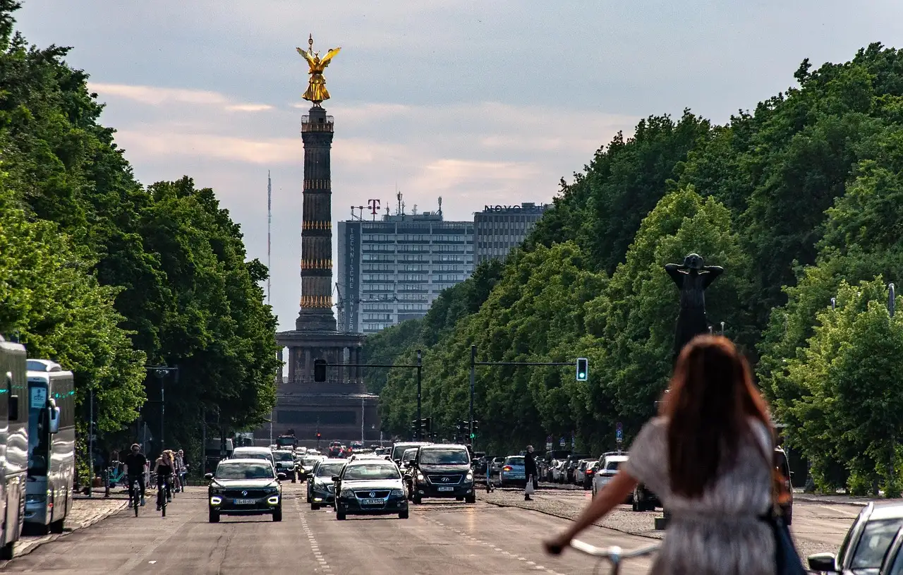 Una mujer va en bici hacia la Columna de la Victoria por una avenida ancha de Berlín con árboles y tráfico