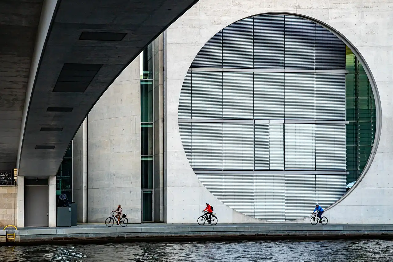 Tres ciclistas junto al Spree pasan ante un edificio gubernamental de hormigón con una enorme ventana redonda