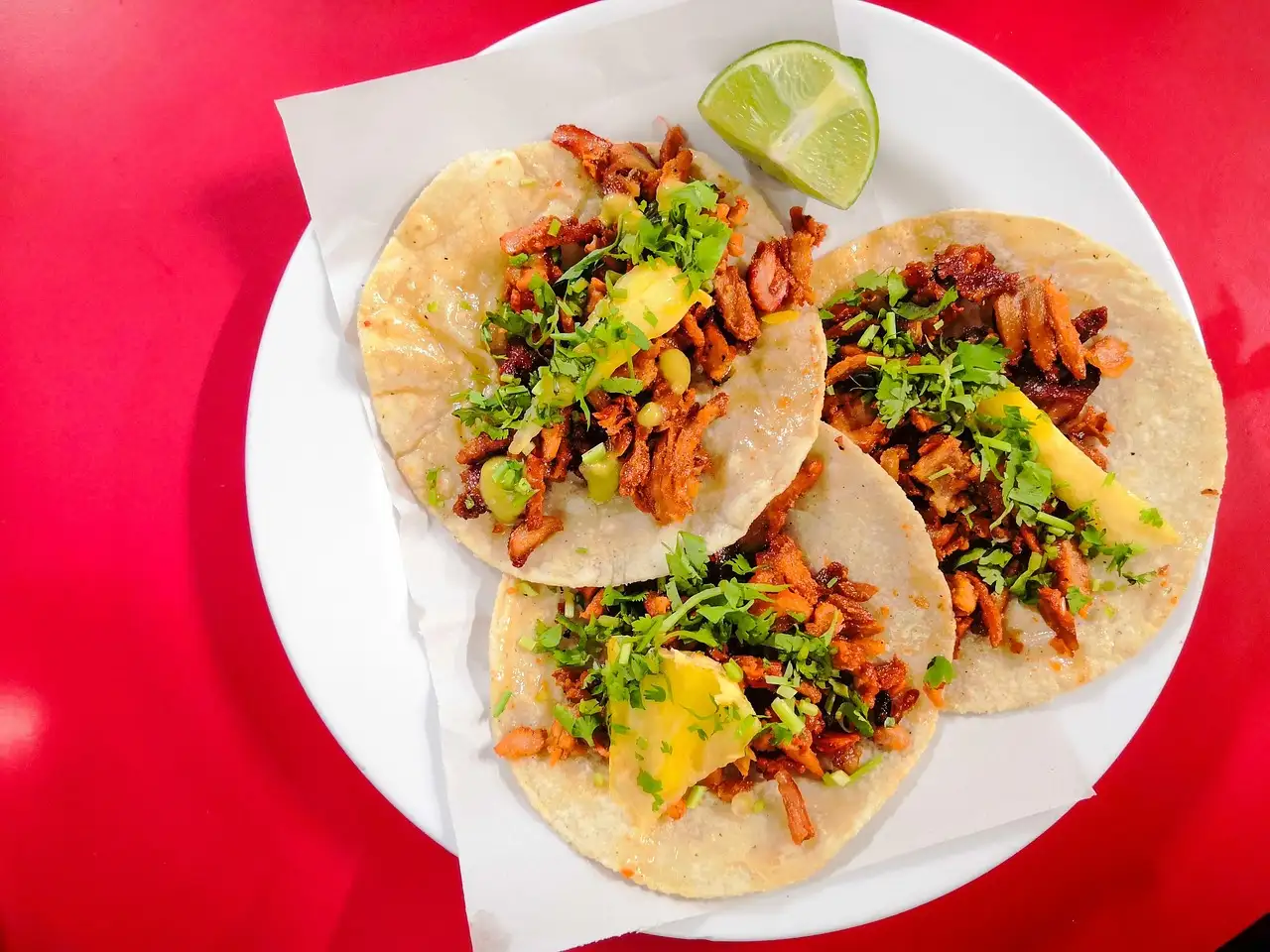 Cuatro tacos blandos con carne, piña y cilantro en un plato blanco, con un gajo de lima