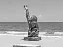 Una estatua de bronce de una familia que se estira hacia el cielo en el malecón de Galveston, con el Golfo detrás, en blanco y negro