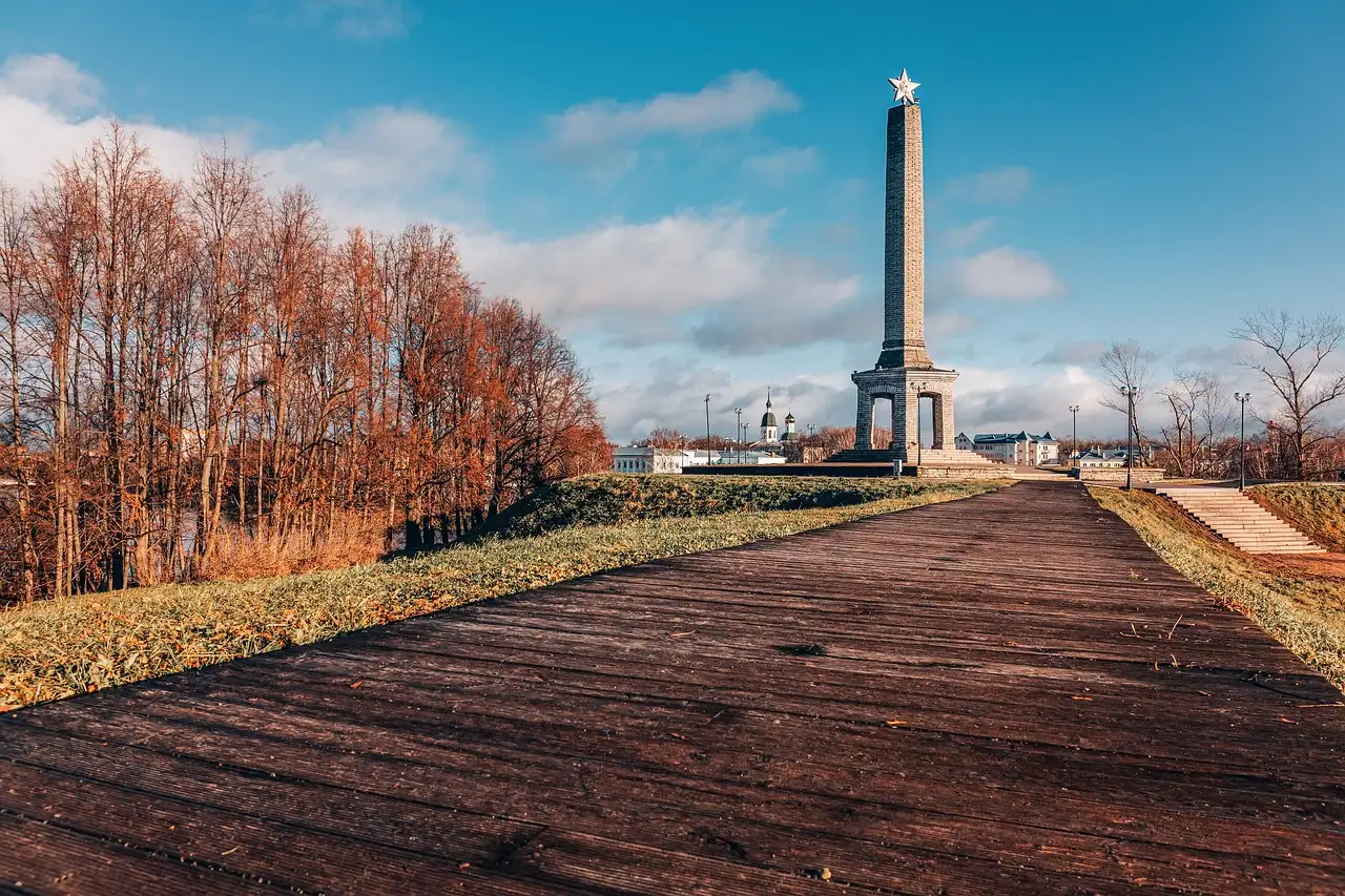 Una alta columna de piedra coronada por una estrella al final de un largo paseo, con árboles otoñales sin hojas