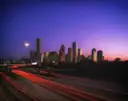 El skyline de Houston al anochecer detrás de una autopista con estelas de luz, bajo un cielo morado y rosa