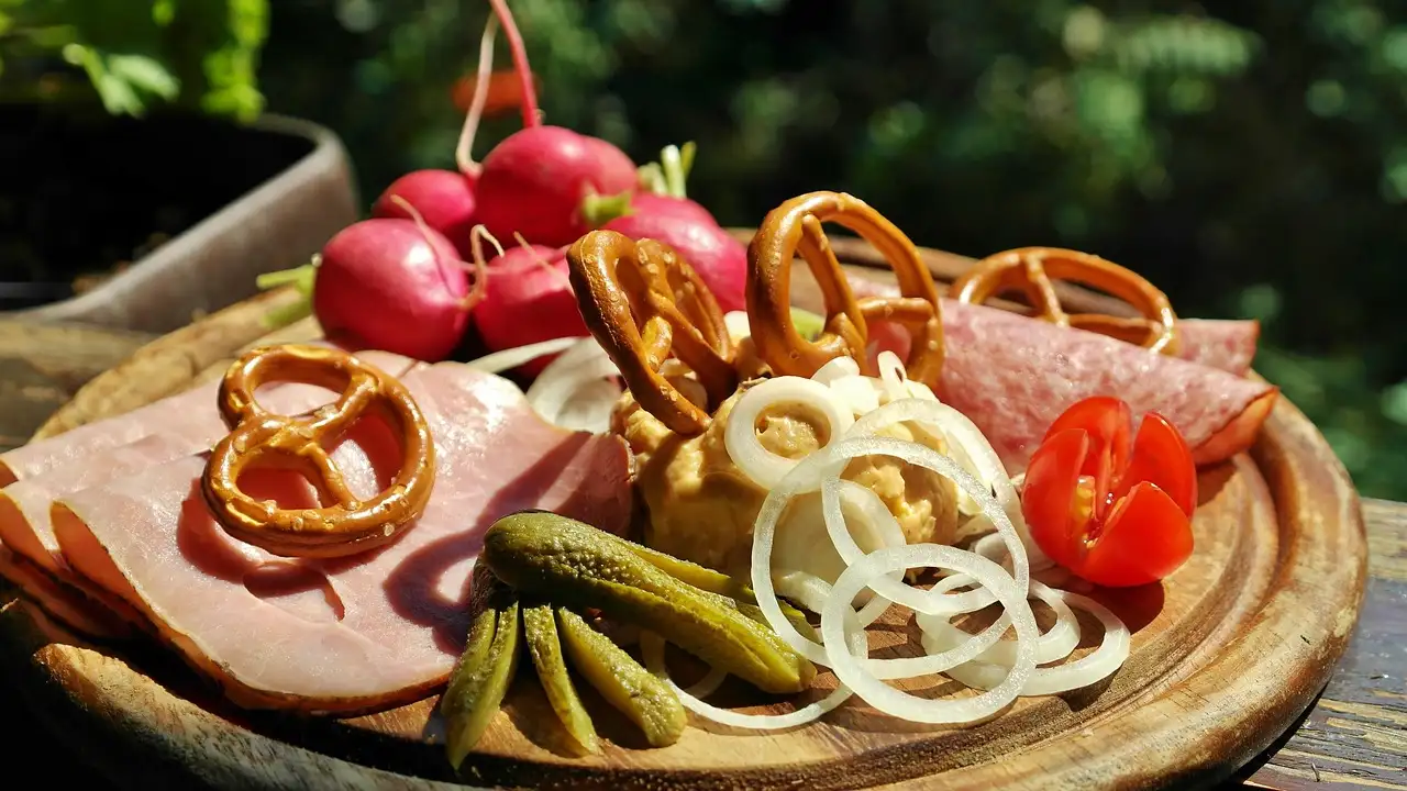Una tabla de madera con jamón, pretzels, rábanos, aros de cebolla, pepinillos y tomates