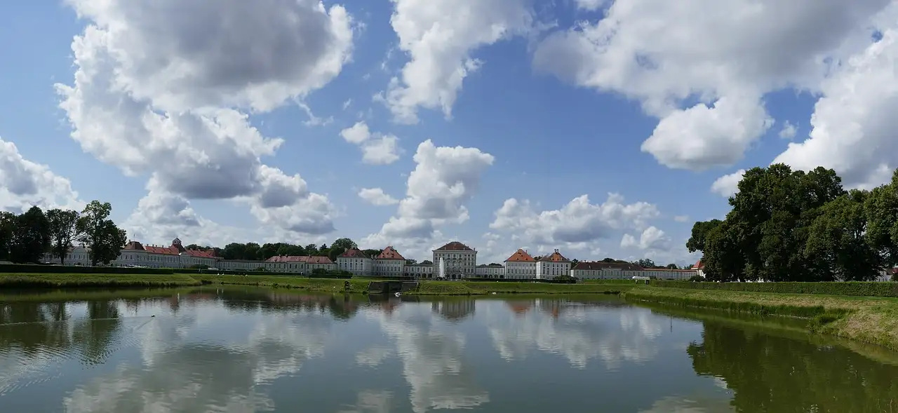 El Palacio de Nymphenburg al otro lado de un lago ancho bajo un cielo azul, con su larga fachada y sus tejados rojos reflejados en el agua
