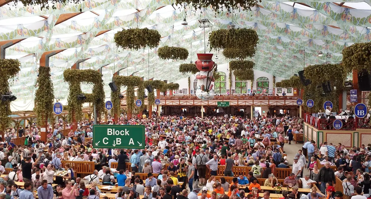 Una carpa del Oktoberfest llena, con largas filas de mesas bajo un techo blanco con guirnaldas verdes