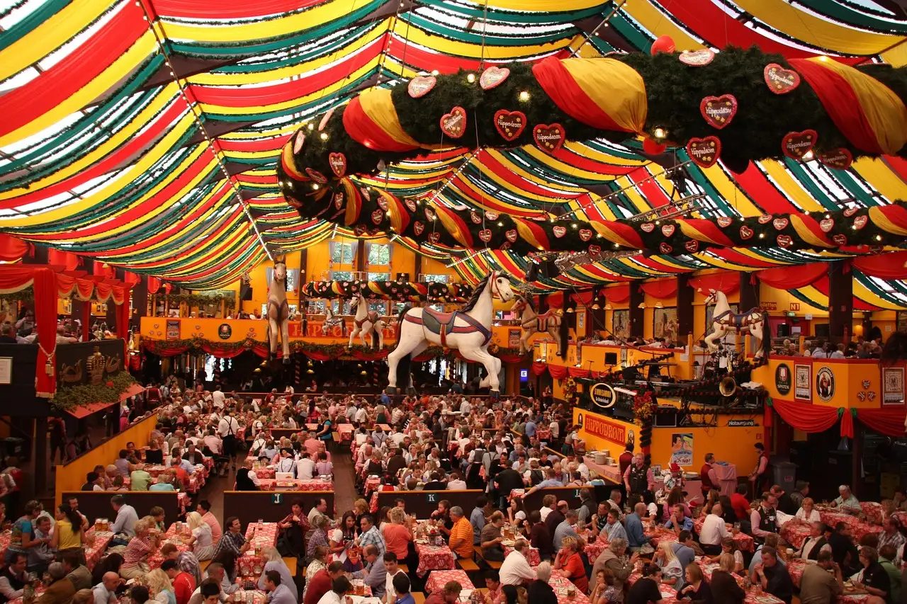 Dentro de una gran carpa de cerveza del Oktoberfest, con mesas largas llenas bajo un techo a rayas rojas, amarillas y verdes