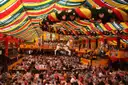 Dentro de una gran carpa de cerveza del Oktoberfest, con mesas largas llenas bajo un techo a rayas rojas, amarillas y verdes