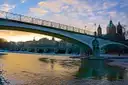 Un puente curvo sobre el río Isar en Múnich al atardecer, con cúpulas de iglesia detrás