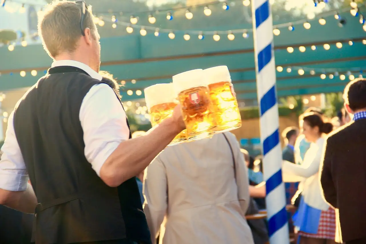 Un camarero con chaleco lleva varias jarras de cerveza llenas por una carpa de la fiesta de Múnich con guirnaldas de luces