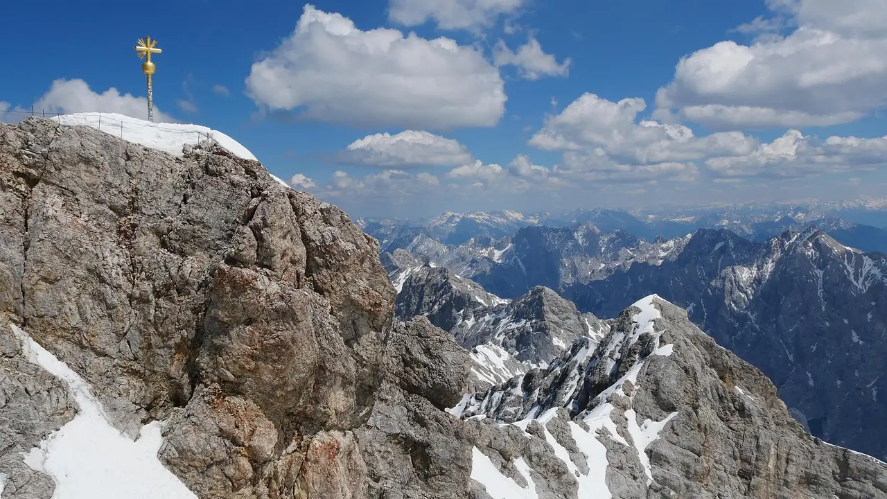 La cima rocosa del Zugspitze con su cruz dorada bajo un cielo azul y manchas de nieve