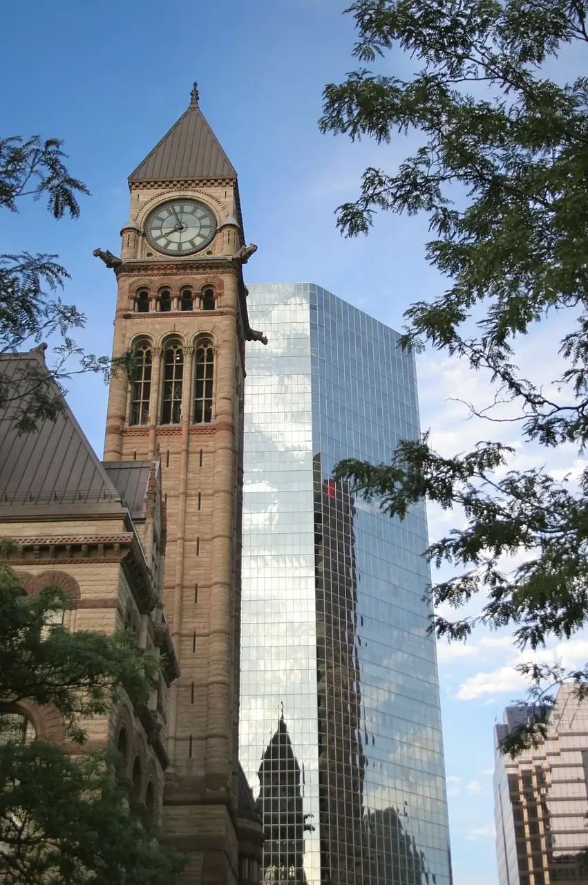 La torre del reloj del antiguo ayuntamiento de Toronto junto a una torre de oficinas de cristal, enmarcada por hojas