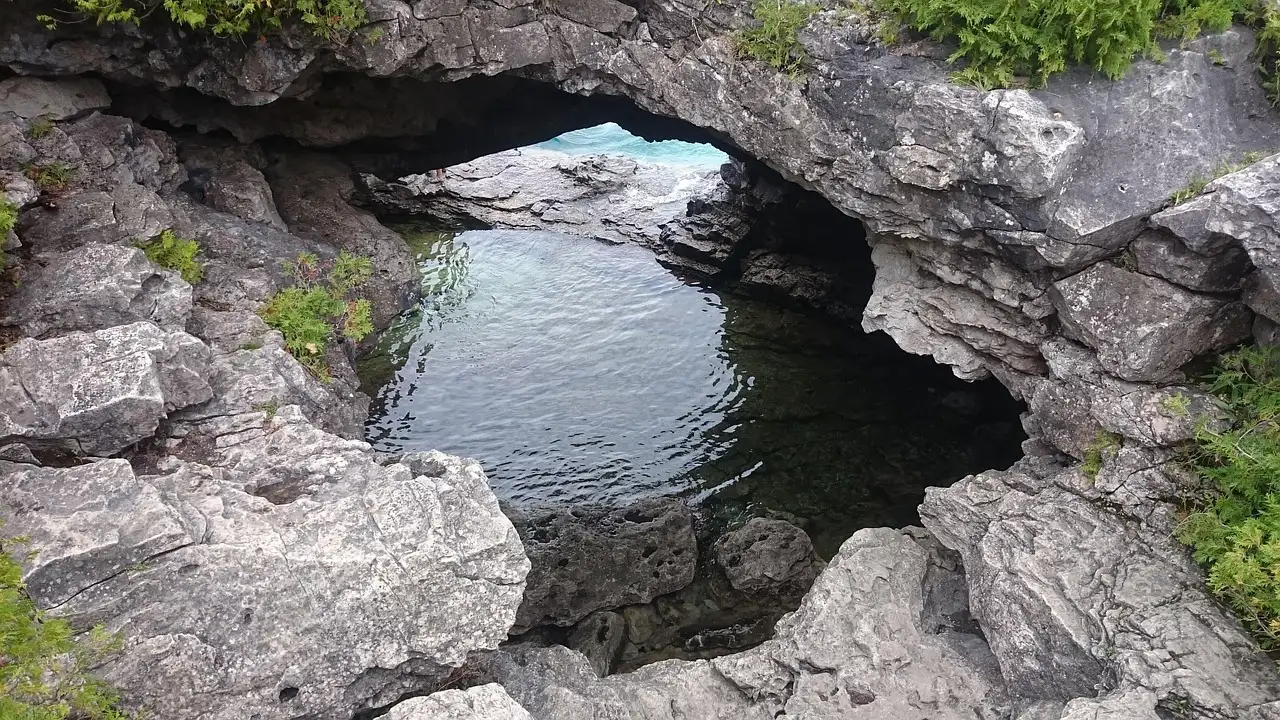 Un arco de cueva de caliza sobre agua transparente, rodeado de roca gris y helechos