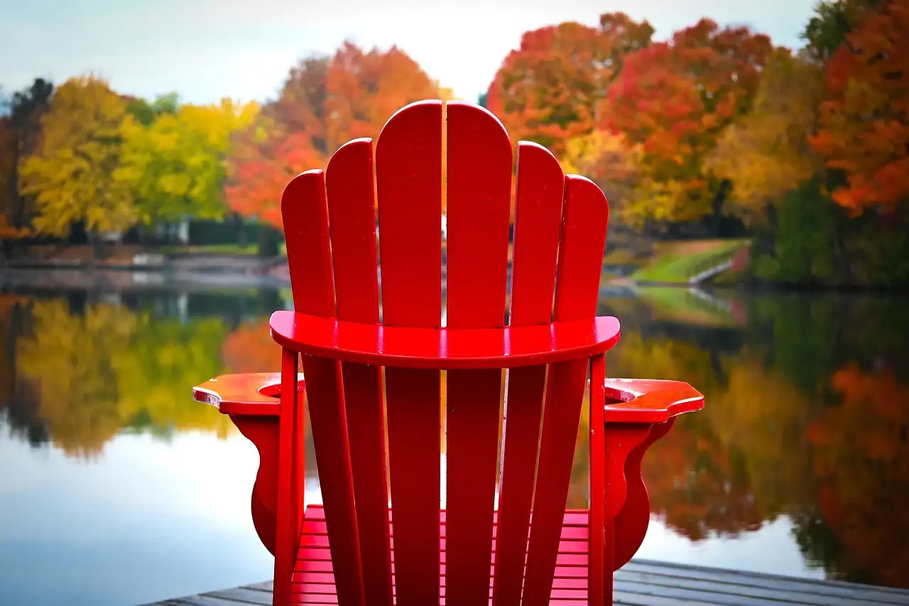 Una silla Muskoka roja en un embarcadero frente a un lago con árboles de otoño