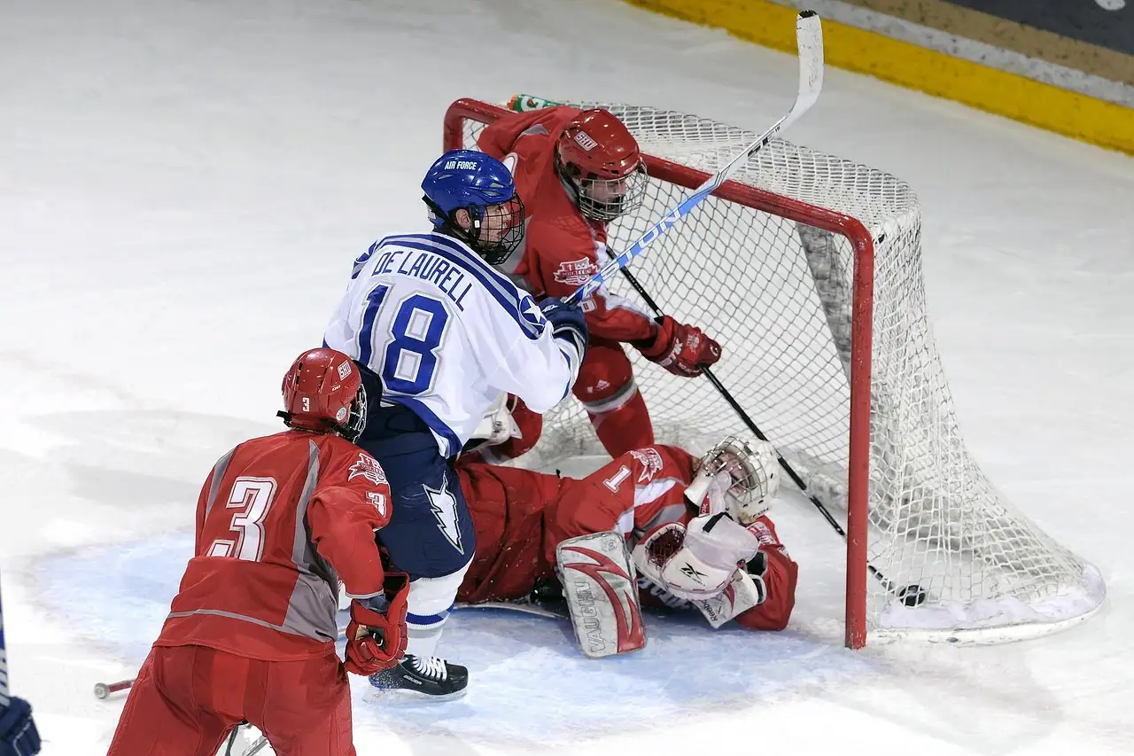 Jugadores de hockey sobre hielo se agolpan delante de una portería roja mientras el portero se lanza al suelo