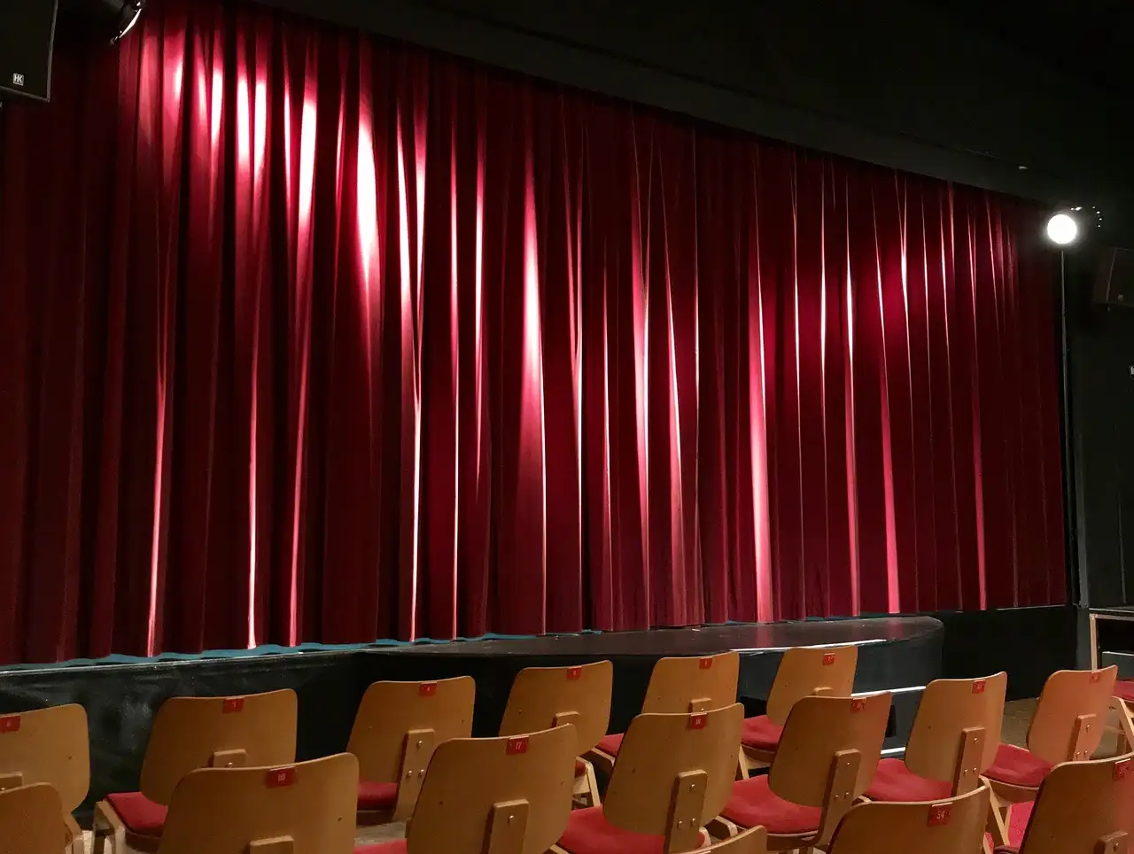 Un telón rojo de teatro bajo una luz cálida, con filas de butacas delante