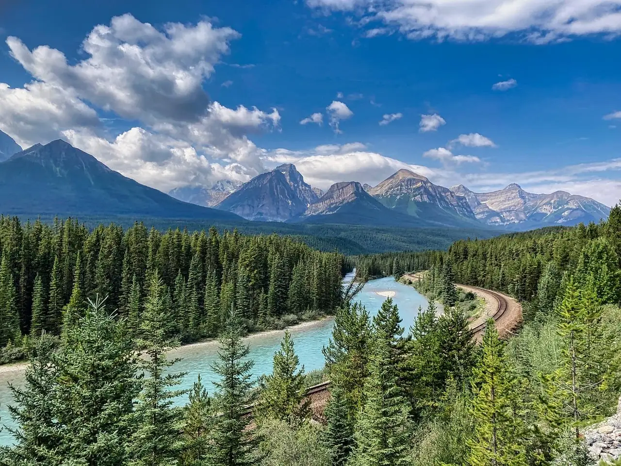 Un río turquesa que serpentea entre pinos bajo las Montañas Rocosas canadienses con manchas de nieve, con una vía de tren que sigue la curva.