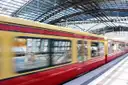 A red and yellow Berlin S-Bahn train speeding past a platform under a glass station roof