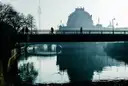 People cross a bridge over the Spree on a hazy morning, with the Bode-Museum dome and the TV Tower behind