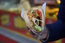 A hand holds a döner kebab in pita bread, wrapped in paper