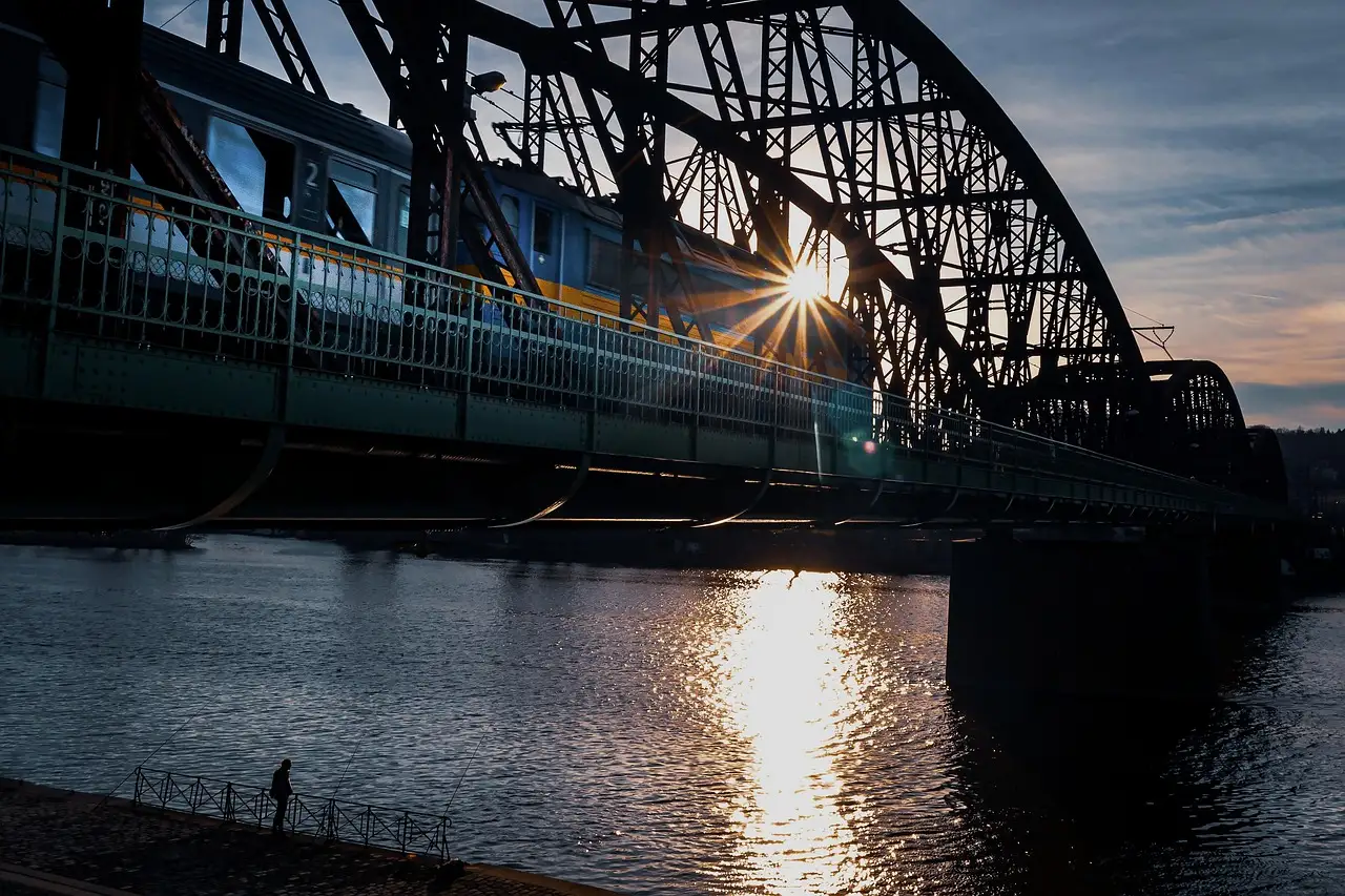 A train crossing a steel arch railway bridge over the Vltava in Prague at sunset