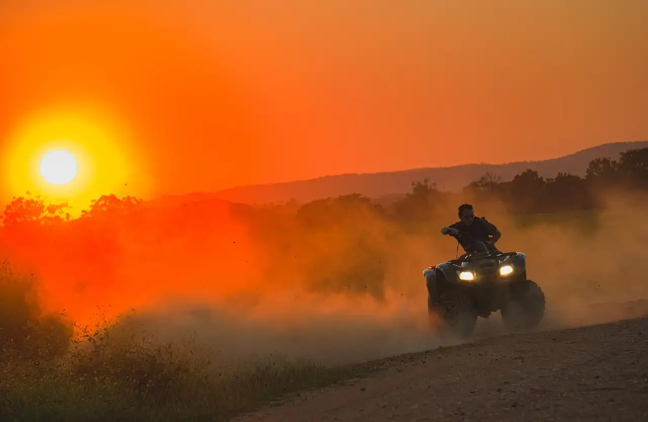 An ATV with its headlights on, riding through dust under an orange evening sky