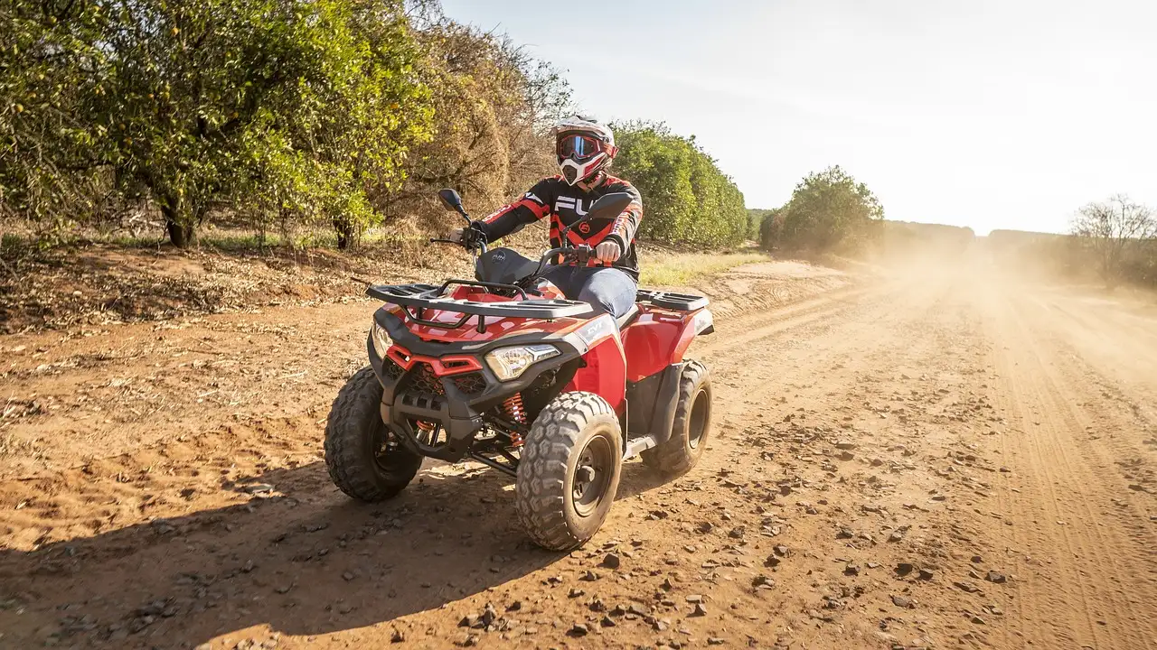 A rider in a helmet on a red ATV kicking up dust on a dirt road between trees