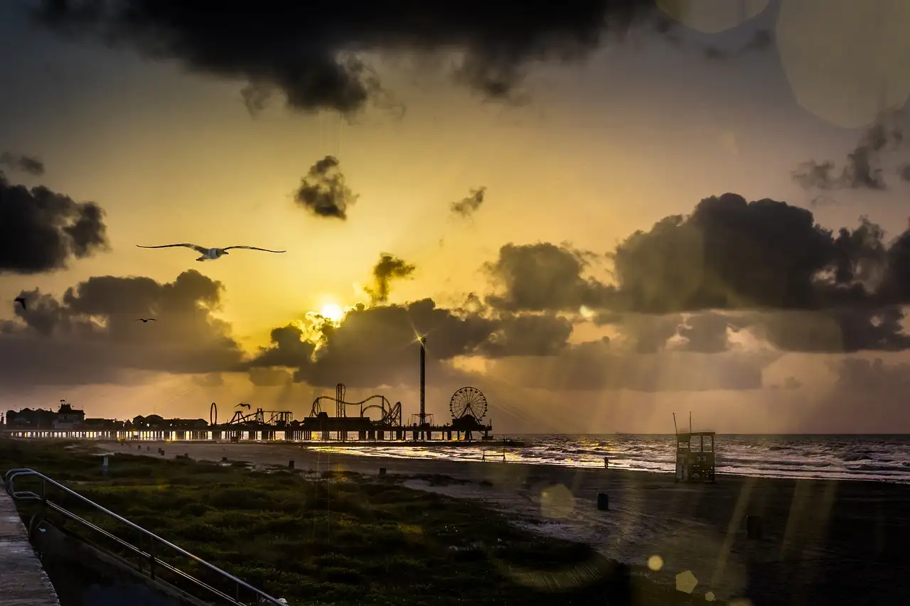 The sun rising over the Gulf behind a long pier with rides at the far end, under dark clouds