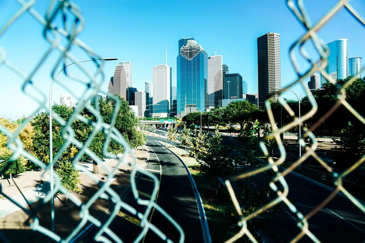 Houston's downtown towers on a clear day seen through a chain-link fence beside a highway