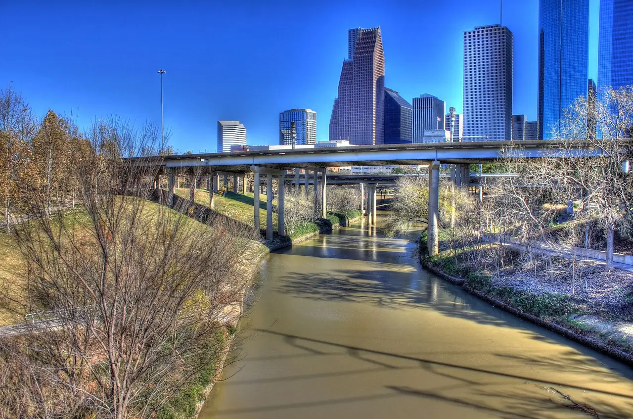 A paved bayou trail with bare trees under a highway overpass and Houston towers behind