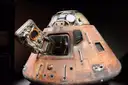 A scorched Apollo command capsule with its hatch open, lit in a dark museum room