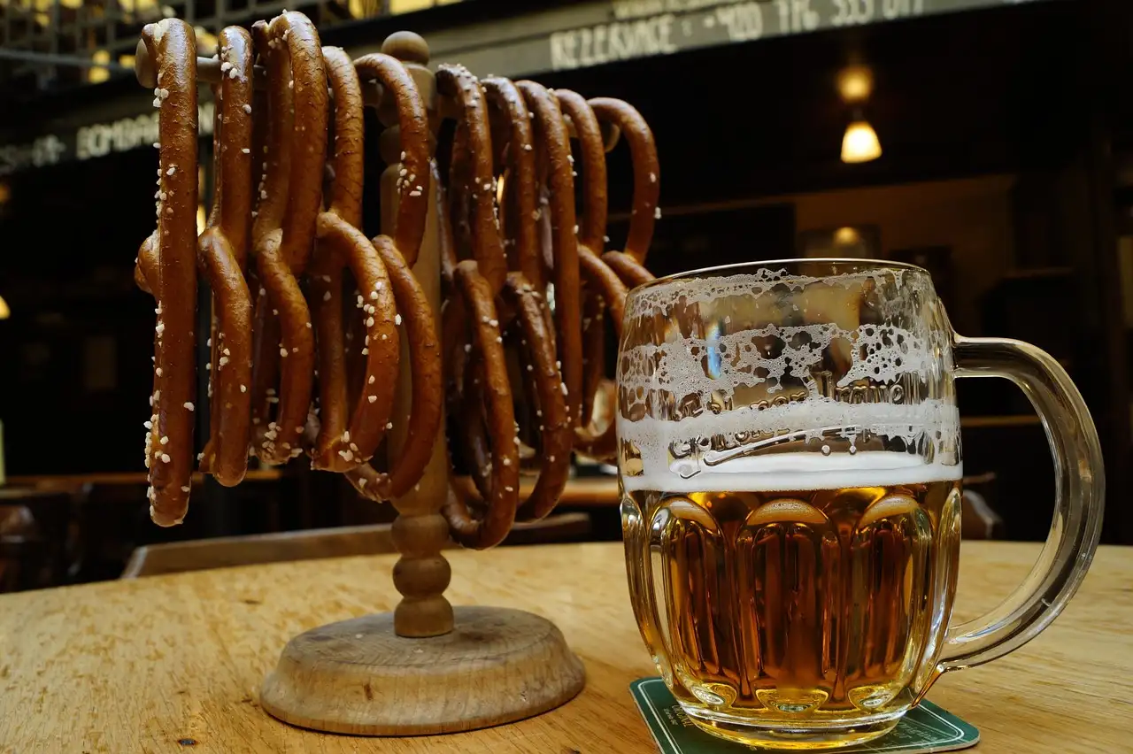 A glass mug of amber beer next to a wooden stand hung with pretzels, in a dim beer hall