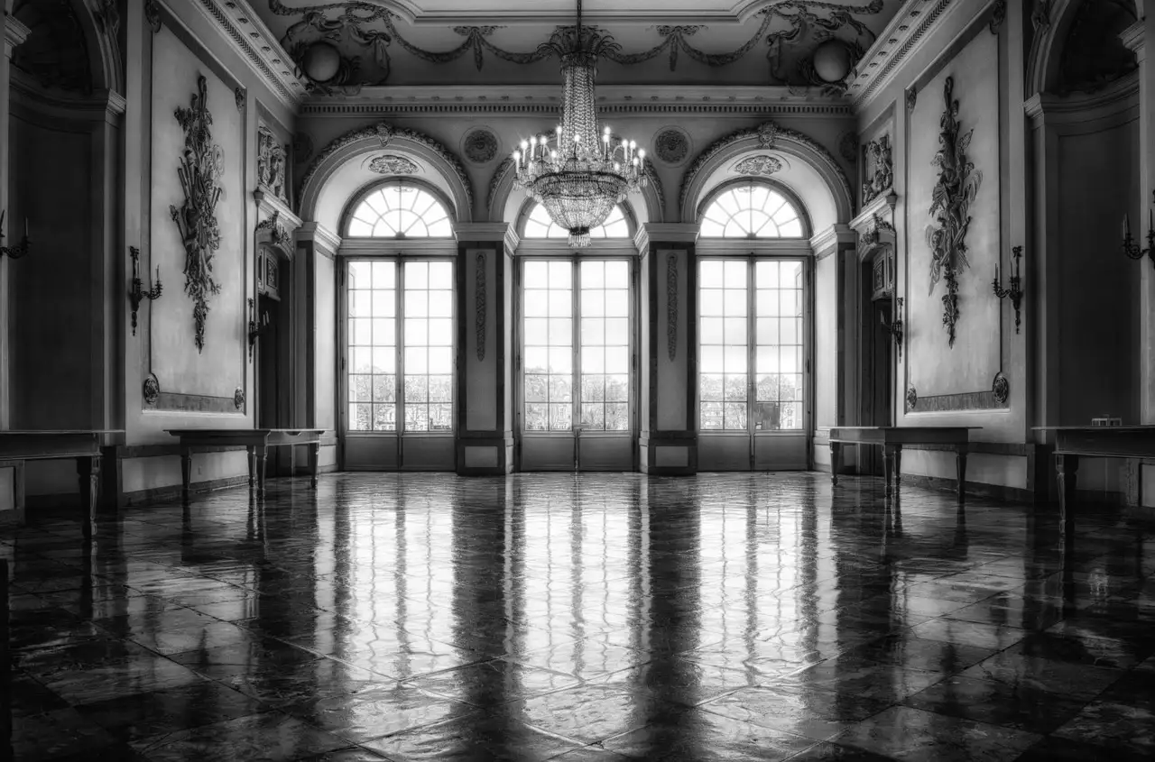 A black-and-white photo of an ornate ballroom with tall windows and a chandelier