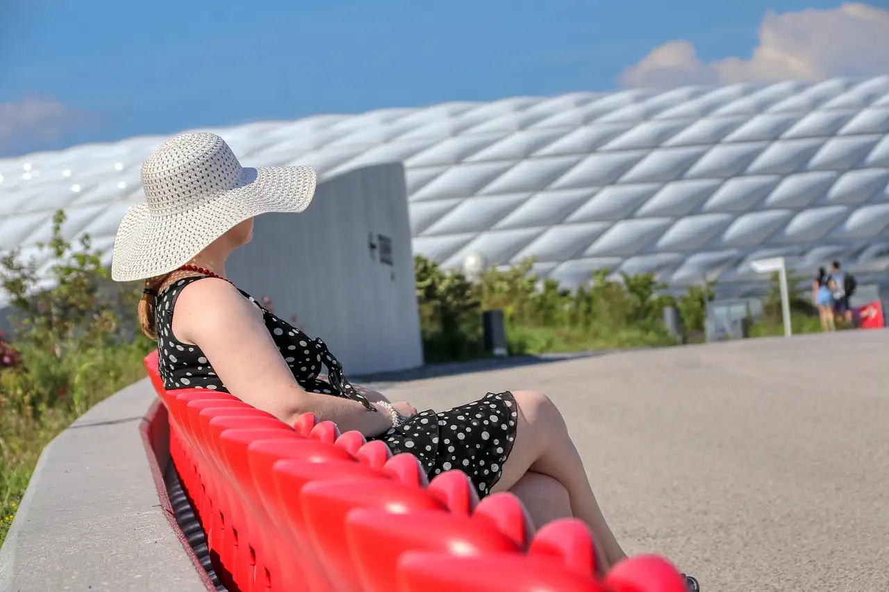 A woman in a sun hat sits on a red bench beside the white cushion-panel facade of the Allianz Arena in Munich