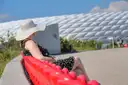 A woman in a sun hat sits on a red bench beside the white cushion-panel facade of the Allianz Arena in Munich