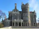 The front of Casa Loma, a grey stone castle with a tall tower and arched windows, under a blue sky