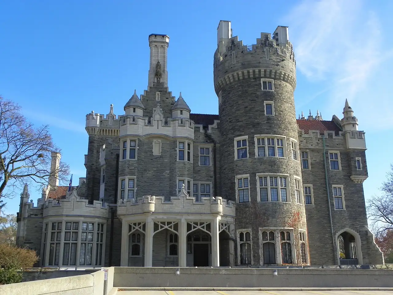 The front of Casa Loma, a grey stone castle with a tall tower and arched windows, under a blue sky