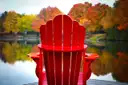 A red Muskoka chair on a dock facing a lake with autumn trees