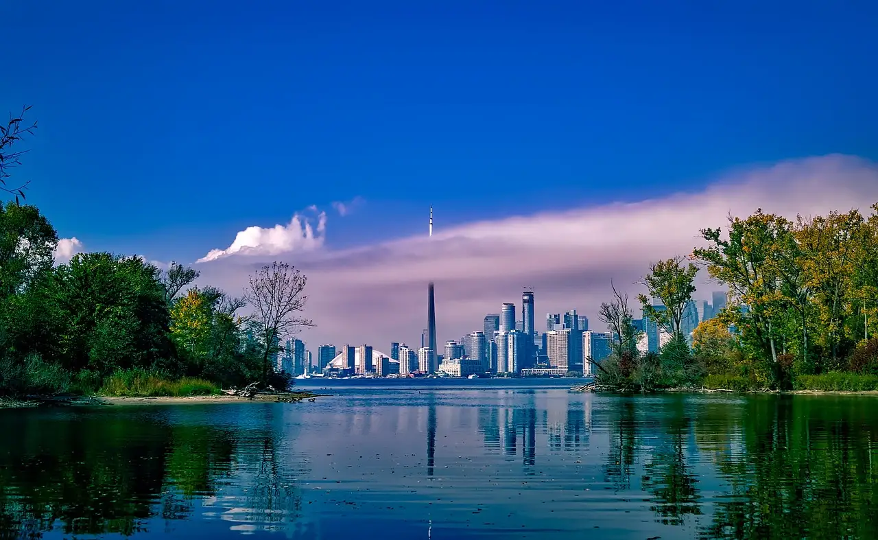 The Toronto skyline and CN Tower across calm water, framed by the green, tree-covered shores of the Toronto Islands.