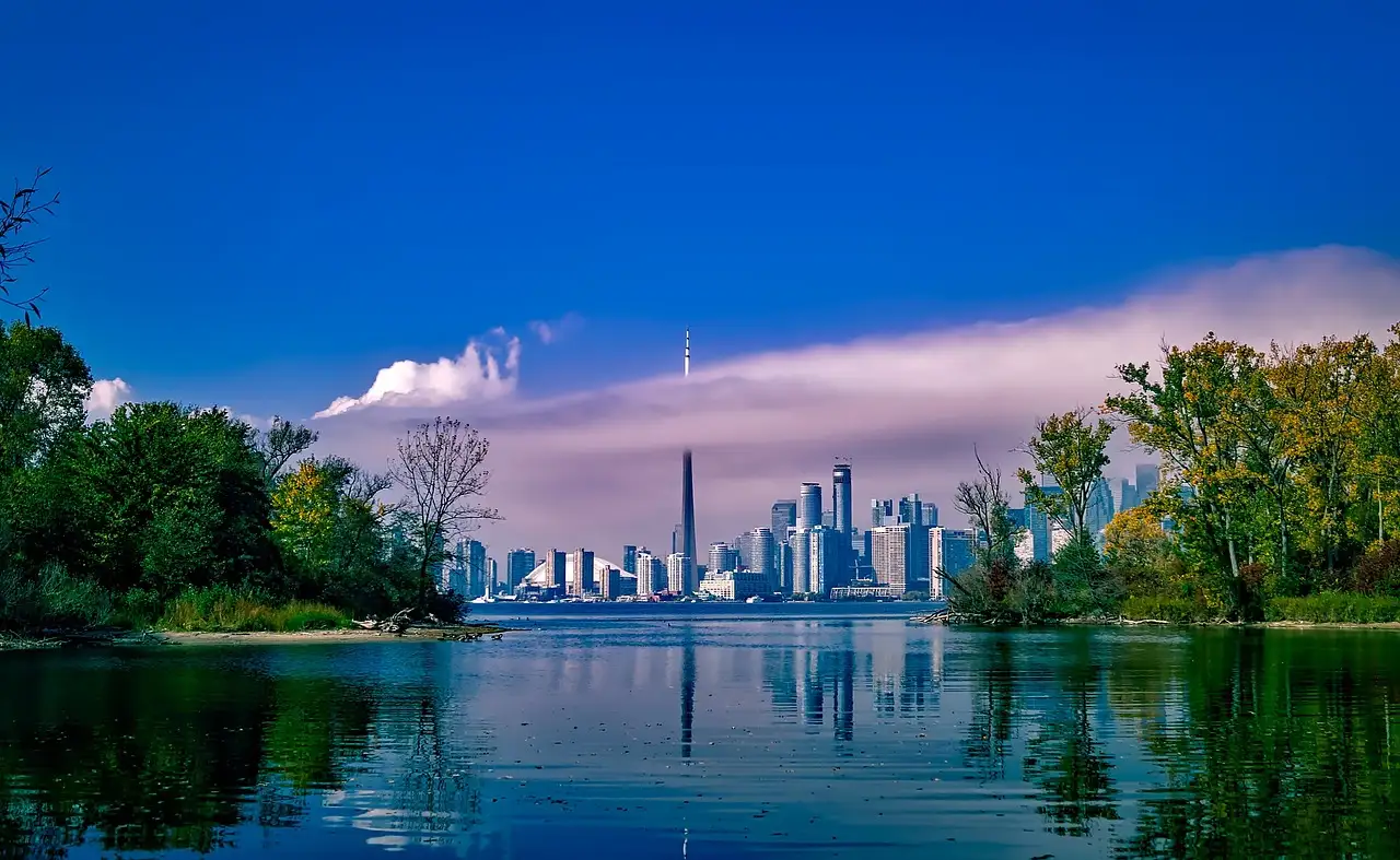 The Toronto skyline and CN Tower across calm water, framed by the green, tree-covered shores of the Toronto Islands.